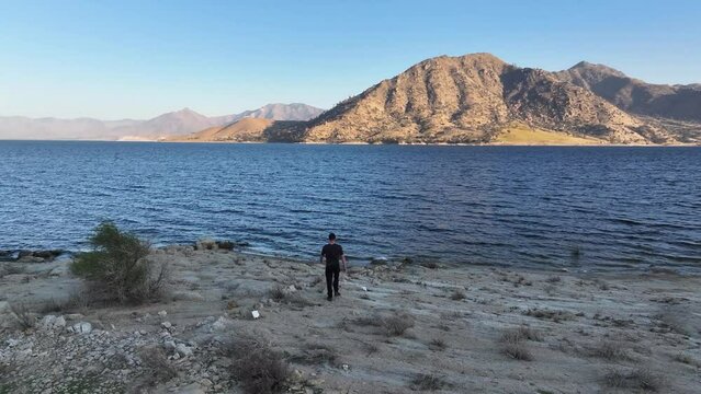 young caucasian male in dark clothing walking up to the waters edge on a rocky outcrop in Lake Isabella Kernville California during sunset 60fps