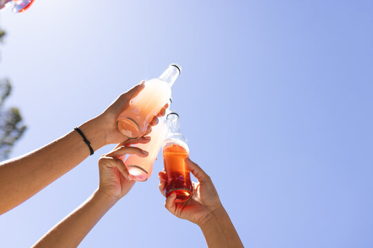 Hands clinking soda bottles against a clear blue sky