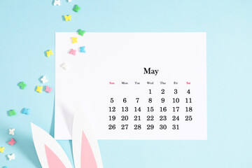 Easter concept. Flat lay, top view of paper desk calendar for May 2024 , easter bunny ears, sprinkles sugar candies on isolated pastel blue background