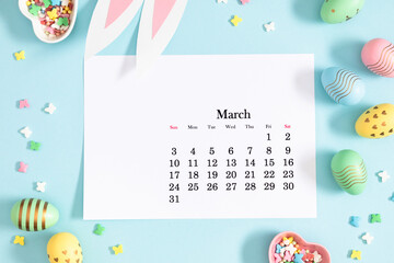 Easter concept. Flat lay, top view of paper desk calendar for March 2024 , easter bunny ears, colorful eggs, sprinkles sugar candies on isolated pastel blue background