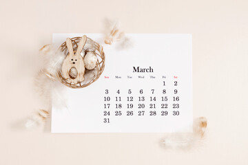 Easter concept. Flat lay, top view of paper desk calendar for March 2024 , Easter eggs, rabbit, feathers in nest on isolated pastel beige background