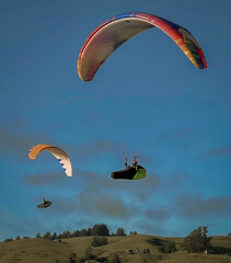Two hang gliders a floating in the air above green hills and with trees. One hand glider is red,...