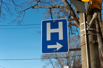 projecting road sign with capital H and arrow pointing left on a blue sky with bare trees in winter © eugen