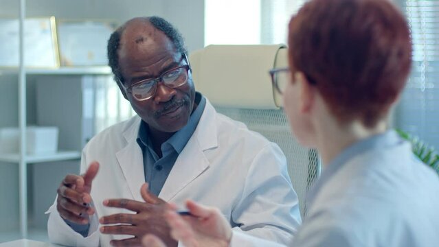 Senior African American general practitioner talking with female colleague about work during meeting in medical office. Over the shoulder view