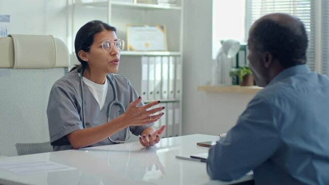 Latin female physician wearing scrubs sitting at table and giving advice about treatment plan to Black patient during medical consultation in clinic