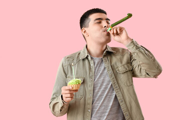 Young man with birthday cake and party whistle on pink background