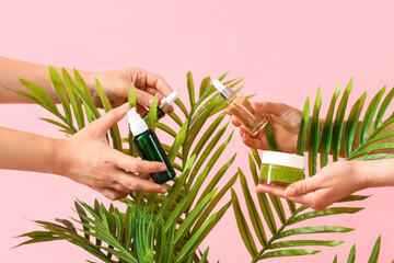 Female hands holding cosmetic products with palm leaves on pink background