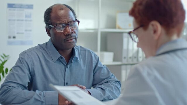 Mature Black man giving medical records to female doctor and complaining about migraine during health consultation in clinic. Over the shoulder view