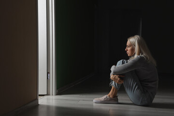 Depressed mature woman sitting in dark room - obrazy, fototapety, plakaty