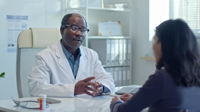 Senior African American doctor sitting behind the table in medical office and discussing symptoms with female patient during health consultation