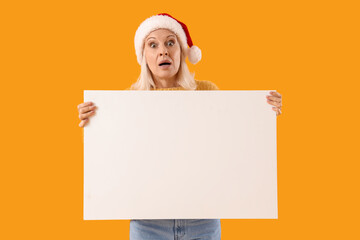 Surprised mature woman in Santa hat holding blank poster on yellow background