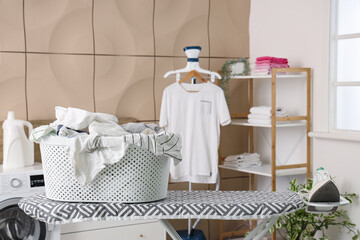 Ironing board with modern iron and basket full of clothes in laundry room