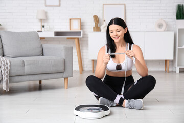 Beautiful young sporty woman with scales and measuring tape sitting at home. Weight loss concept