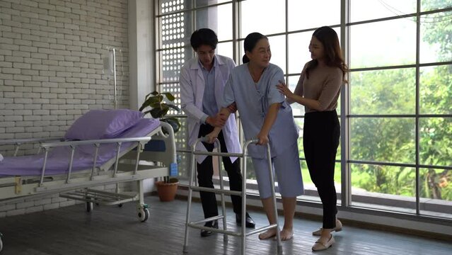 A male physical therapist with an Asian relative works with an elderly woman to walk using a walker in a rehab center or nursing home/hospital.