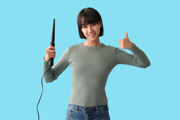 Beautiful young happy woman with curling iron showing thumb-up gesture on blue background