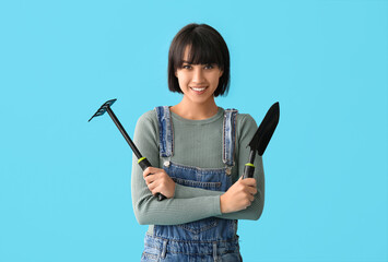 Beautiful young happy female gardener with gardening rake and shovel on blue background