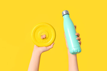 Female hands with barbell plate and bottle of water on yellow background, closeup