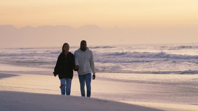 Full length shot of casually dressed loving young couple walking hand in hand along sandy shoreline watching beautiful sunrise morning over beach and sea in South Africa - shot in slow motion