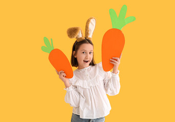 Shocked little girl in Easter bunny ears with paper carrots on yellow background