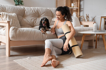 Sporty young African-American woman with cocker spaniel and yoga mat at home
