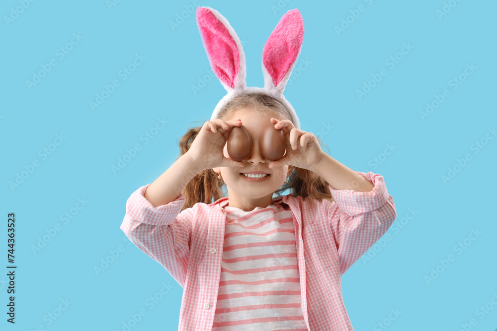 Wall mural Cute little girl in bunny ears with chocolate Easter eggs on blue background