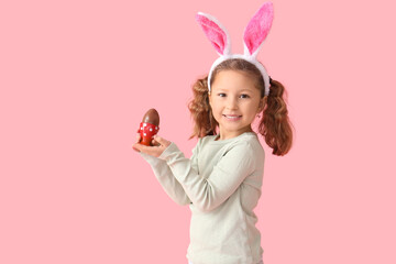 Cute little girl in bunny ears with chocolate Easter egg on pink background