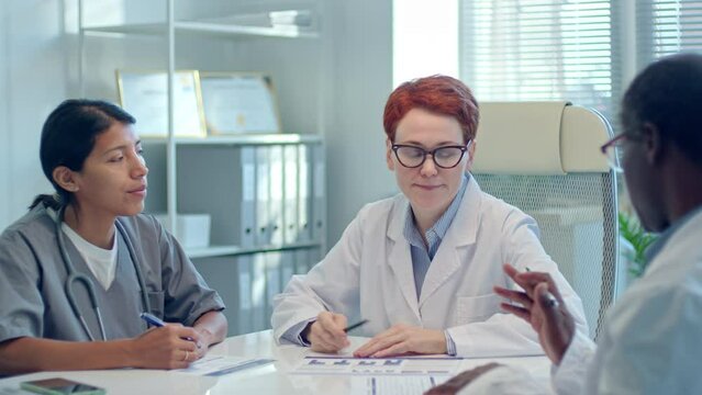 Group of multiracial doctors sitting around meeting table and discussing documents during consilium in medical office