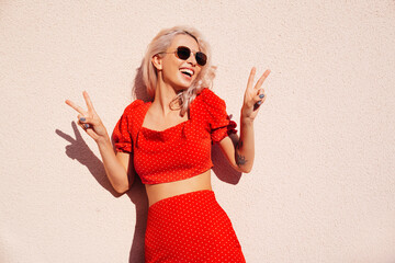 Young beautiful smiling hipster woman in trendy summer red top and skirt clothes. Carefree female posing in street. Positive model at sunset. Cheerful and happy. In sunglasses. Shows peace sign