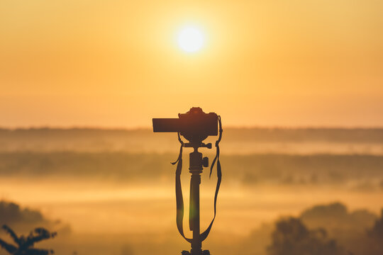 A professional photographer's camera on a tripod looks at the sunrise. Countryside landscape with camera on tripod, sunrise and morning fog.