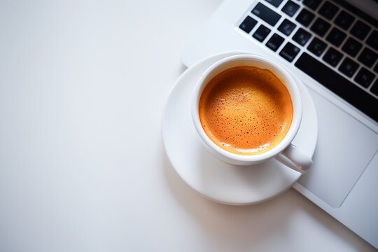 A simple and clean image of a cup of coffee next to a laptop on a white desk, perfect for concepts related to work and productivity.