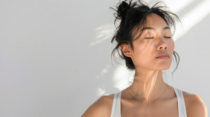 Beautiful and stylish young Asian woman in white tank top with her eyes closed, meditate and contemplate in white room in sunlight from window