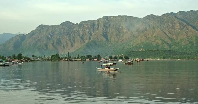 Wide view of Dal Lake in Srinagar, Kashmir, India, featuring traditional Shikara boats, floating houseboats, calm water reflections, and the majestic Himalayan mountains in the background.