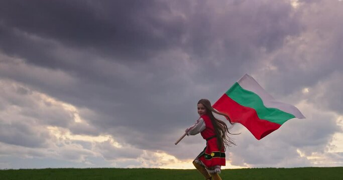 Bulgarian girl standard bearer in ethnic folklore costume with bulgarian flag running in green field and celebrating 3th March Liberation day of Bulgaria
