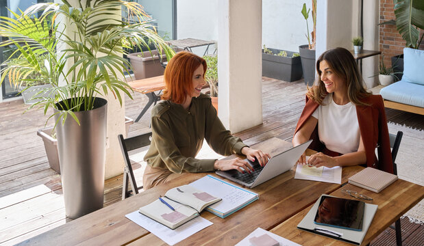 Two happy business women of young and middle age talking in green cozy office sitting at desk. Professional ladies executives having conversation using laptop at work. Authentic shot, top view.