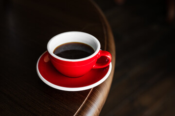 A red cup of black coffee on a saucer on the edge of wooden table, copy space