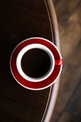 Top view of a red cup of black coffee on a saucer on the edge of wooden table, copy space - 744463910