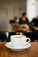 A white cup of black coffee on a saucer on the wooden table in a cafe, people in bokeh - 744464924
