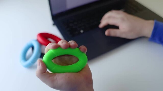 Hand gripper. Man squeezes rubber expander while working on his laptop. Concept of combining useful activity with necessary work. Training hand and arm muscles. Multi-colored mini training apparatus