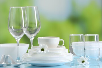 Set of many clean dishware and glasses on light blue table against blurred green background