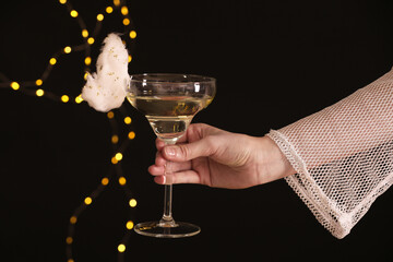 Woman holding glass of cocktail decorated with tasty cotton candy on black background with blurred lights, closeup