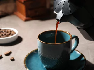 Tuinposter Pouring aromatic coffee from moka pot into cup at light table, closeup © New Africa