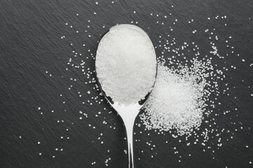 Granulated sugar and metal spoon on black table, top view