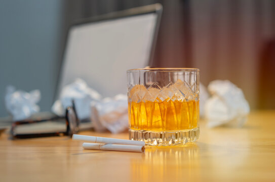 Glass whiskey with ice cubes and cigarettes on wooden desk with blurred image of scraps of paper
