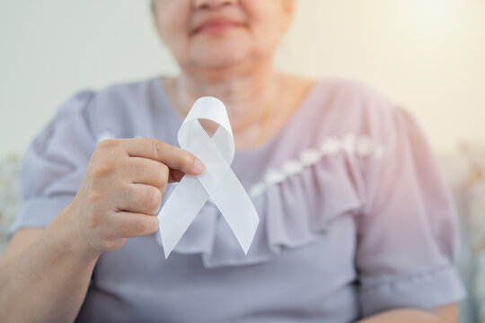Selective focus of older female hand is holding a white ribbon sit at home background. A symbol of the fight against lung cancer.