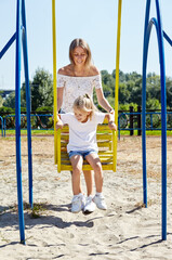 Mother with daughter on swing on a playground in the summer city park. Childhood, leisure and people concept - happy family rest and have a good time