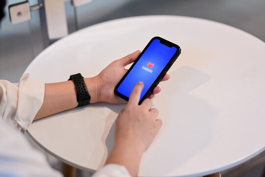 Chiang Mai, Thailand - Feb 24, 2024: A woman uses the Lazada application on her iPhone while sitting at a table in a cafe. The Lazada logo is displayed on the smartphone screen.