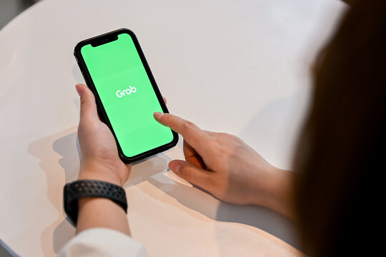 Chiang Mai, Thailand - Feb 24, 2024: A woman uses the Grab application on her iPhone while sitting at a table in a cafe. The Grab logo is displayed on the smartphone screen.