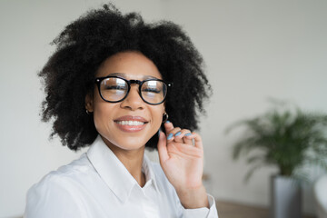 Confident woman with glasses holds earpiece, smiling in a minimalist room.