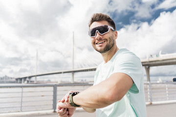 Smiling man with sporty sunglasses checks smartwatch by sunlit bridge.