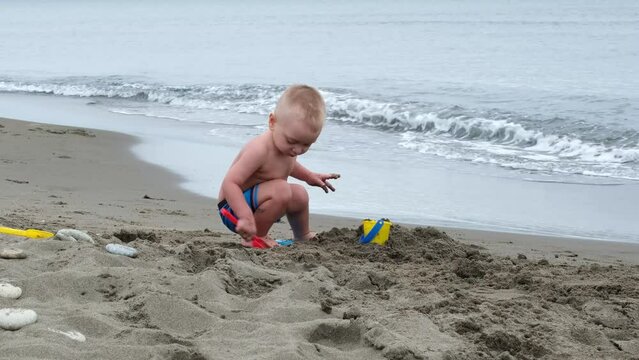A 3-year-old boy with blond hair enthusiastically plays on the beach, pours sand into a bucket with a shovel.